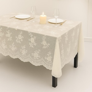 May include: A beige rectangular tablecloth with a floral lace pattern and scalloped edges. The table is set with white plates, wine glasses, a lit candle, and gold-colored cutlery. The table is supported by black legs.