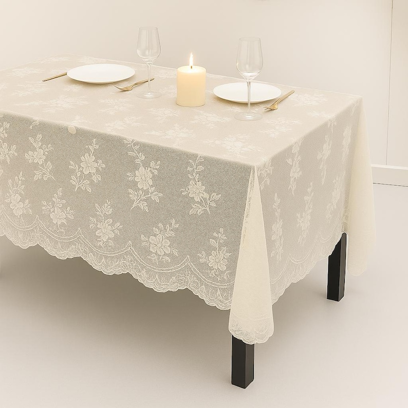May include: A beige rectangular tablecloth with a floral lace pattern and scalloped edges. The table is set with white plates, wine glasses, a lit candle, and gold-colored cutlery. The table is supported by black legs.