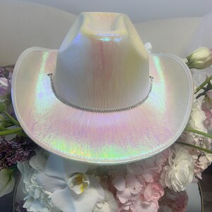 May include: A white iridescent cowboy hat with a silver rhinestone trim. The hat is sitting on a bed of white and pink flowers.