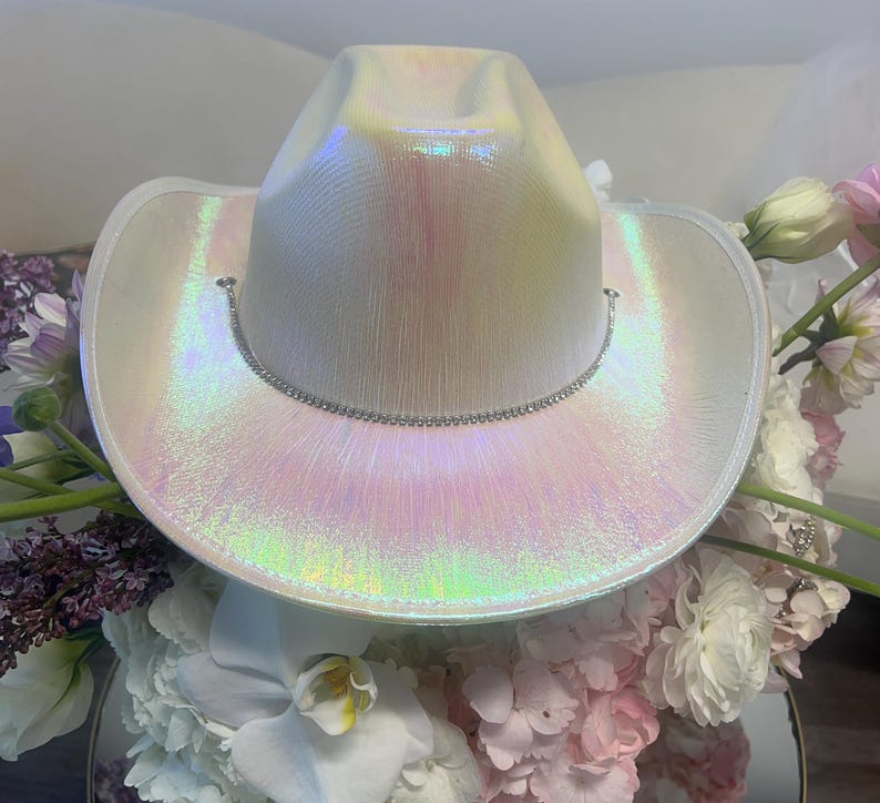 May include: A white iridescent cowboy hat with a silver rhinestone trim. The hat is sitting on a bed of white and pink flowers.