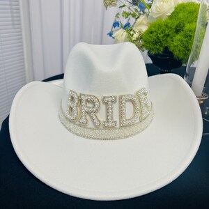 May include: White cowboy hat with a pearl and rhinestone embellishment that reads "BRIDE".