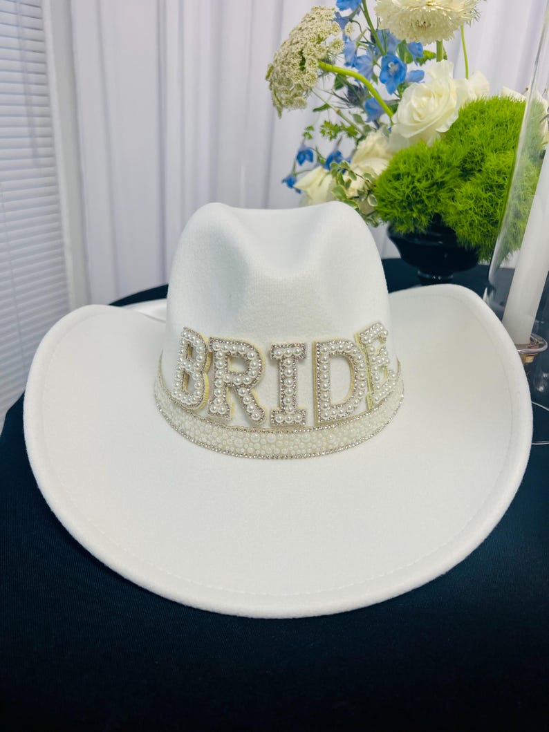 May include: White cowboy hat with a pearl and rhinestone embellishment that reads "BRIDE".
