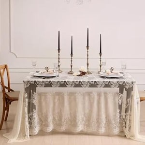 May include: A dining table set for a formal occasion. The table is covered with a gray tablecloth with white lace trim and a matching lace skirt. Black candles in ornate holders, plates, and glasses complete the elegant setting.