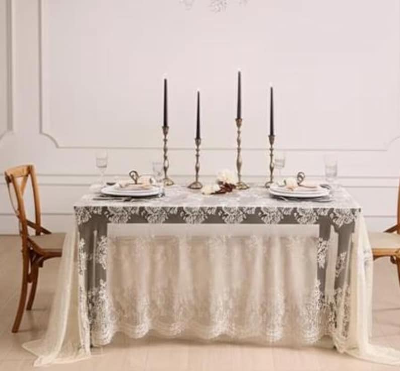 May include: A dining table set for a formal occasion. The table is covered with a gray tablecloth with white lace trim and a matching lace skirt. Black candles in ornate holders, plates, and glasses complete the elegant setting.