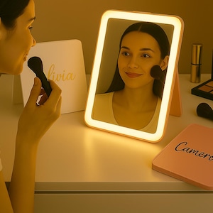 May include: A lighted makeup mirror with a rectangular shape and a pink frame. A woman is applying makeup with a brush. A compact with the name "Cameron" is visible, along with other makeup items.