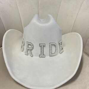 May include: White cowboy hat with the word "BRIDE" spelled out in pearl-embellished letters.