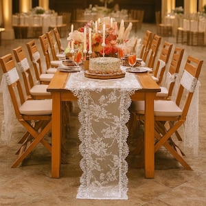 Peut inclure: Une longue table en bois dressée pour un événement spécial, ornée d'un chemin de table en dentelle blanche et d'une pièce maîtresse florale. Des chaises en bois avec des coussins crème sont disposées autour de la table. Un gâteau et des boissons sont posés sur la table, avec des bougies et des fleurs.