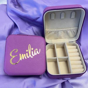 Pode incluir: Caixa de joias roxa com uma inscri&ccedil;&atilde;o dourada que diz "Emilia". A caixa tem uma tampa com dobradi&ccedil;as e um compartimento para an&eacute;is e brincos.