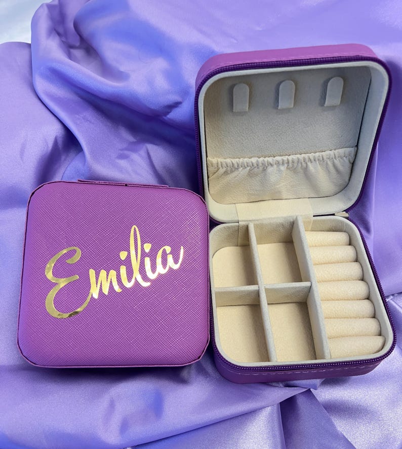 Pode incluir: Caixa de joias roxa com uma inscri&ccedil;&atilde;o dourada que diz "Emilia". A caixa tem uma tampa com dobradi&ccedil;as e um compartimento para an&eacute;is e brincos.