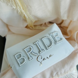 Peut inclure: Pochette de mariée blanche avec le mot "BRIDE" écrit en perles. Le sac a une inscription dorée qui dit "Sara".