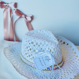 May include: White crochet cowboy hat with a gold and iridescent sequin trim. A white tag is attached to the hat with the text "Ella Will you be my bridesmaid?"