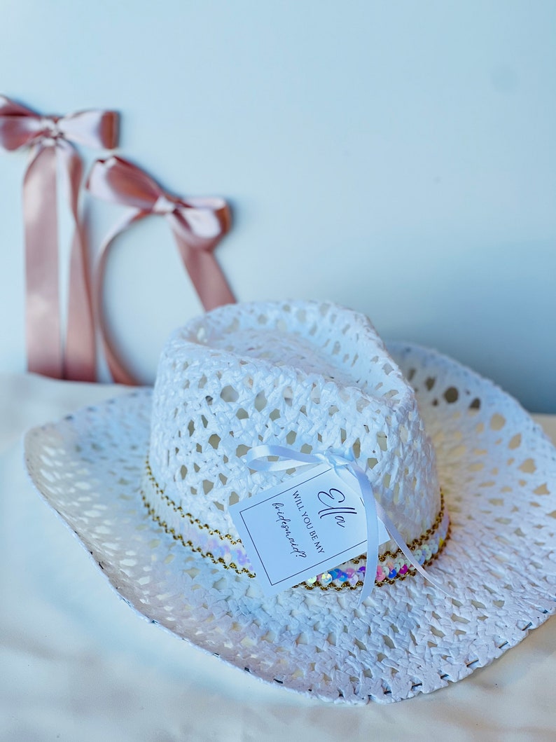 May include: White crochet cowboy hat with a gold and iridescent sequin trim. A white tag is attached to the hat with the text "Ella Will you be my bridesmaid?"