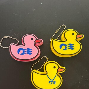 Op de afbeelding: Drie rubberen eend-sleutelhangers met metalen kogelkettingen. Eén is roze met een blauw golflogo, één is geel met een blauw golflogo en één is geel met een blauw drietand-achtig symbool. Elke eend heeft een zwarte omlijning en een oranje snavel.