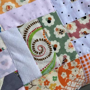 May include: A patchwork quilt featuring a variety of fabric squares with diverse patterns including orange and green floral prints, white fabric with black polka dots, and a central square displaying a brown and green spiral design on a cream background.