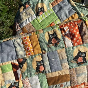 May include: A patchwork quilt featuring owl designs. The quilt is composed of various fabric squares in shades of green, orange, brown, and gray. Each square is stitched together, creating a textured surface. The owl motifs are set against a moonlit background.