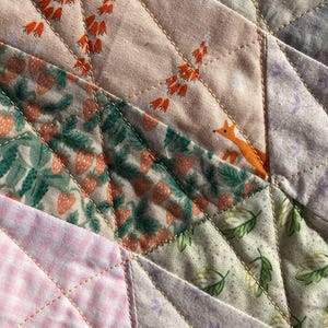 May include: Close-up of a patchwork quilt with various fabric patterns. The quilt features pink, green, and lavender fabrics with floral and geometric designs. Orange foxes and floral patterns are visible. The quilt is stitched with gold thread.