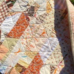 May include: A patchwork quilt with a geometric design. The quilt features various fabrics in shades of white, brown, orange, and beige. Some patches have floral and animal prints. The quilt's edge is a light pink with a repeating pattern.