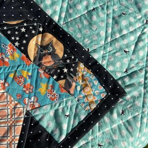 May include: A patchwork quilt with a black border and a variety of patterned fabrics. The quilt features a central panel with an owl in front of a full moon, surrounded by stars. Other panels have floral and geometric designs, and the background is a light teal with white polka dots.