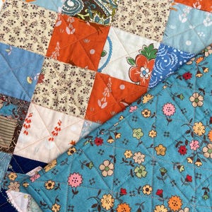 May include: A patchwork quilt featuring a variety of fabric squares in shades of orange, blue, and cream. The quilt displays diverse patterns including floral prints, paisley, and small geometric motifs, all joined by a diamond-shaped stitch pattern.