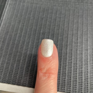 May include: A close-up of a finger with a light, shimmery, pearlescent nail polish. The nail is square-shaped and reflects light, creating a subtle sparkle. The skin tone is fair, and the background is a textured grey surface.