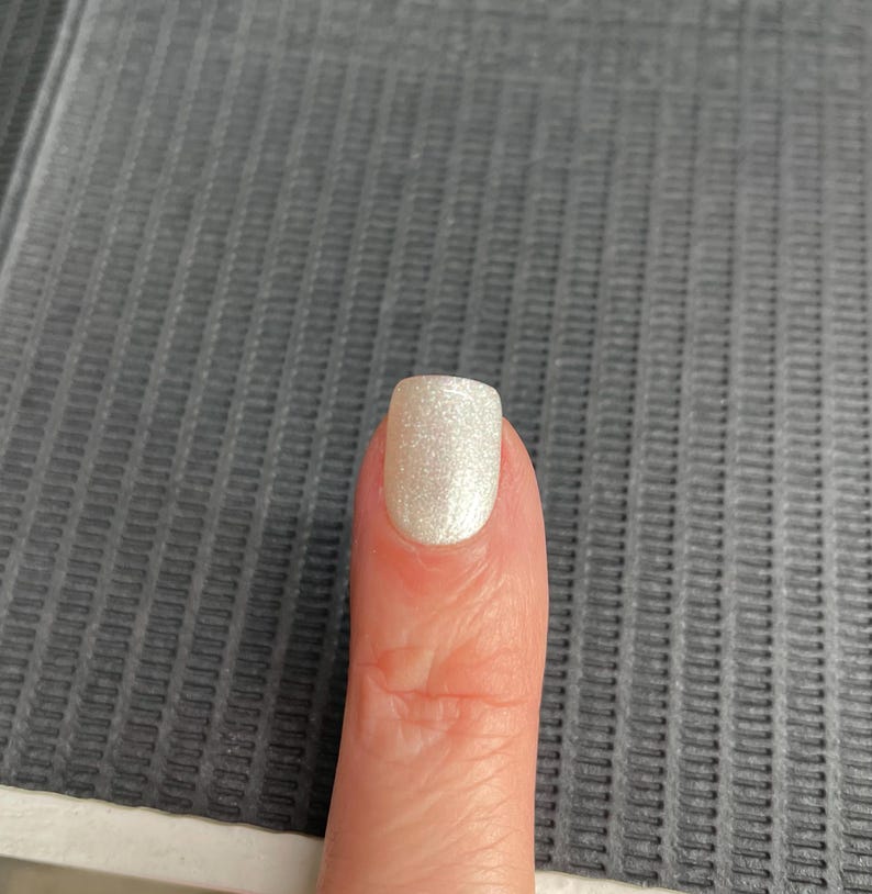May include: A close-up of a finger with a light, shimmery, pearlescent nail polish. The nail is square-shaped and reflects light, creating a subtle sparkle. The skin tone is fair, and the background is a textured grey surface.