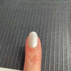 May include: A close-up of a finger with a polished nail. The nail is painted with a pearlescent, shimmering white colour. The nail has an oval shape and reflects light, creating a subtle glow.