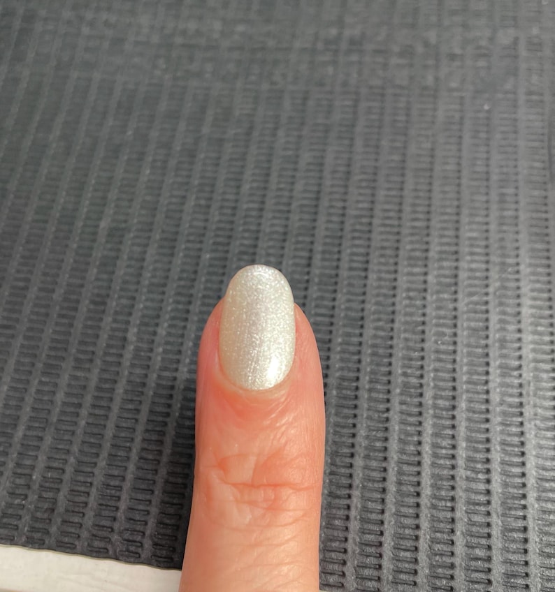 May include: A close-up of a finger with a polished nail. The nail is painted with a pearlescent, shimmering white colour. The nail has an oval shape and reflects light, creating a subtle glow.