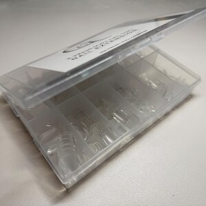 May include: A clear plastic box with a hinged lid, divided into multiple compartments. The box contains numerous clear, artificial nail tips in various sizes. The lid has a white label with black text. The box is open, revealing the contents.