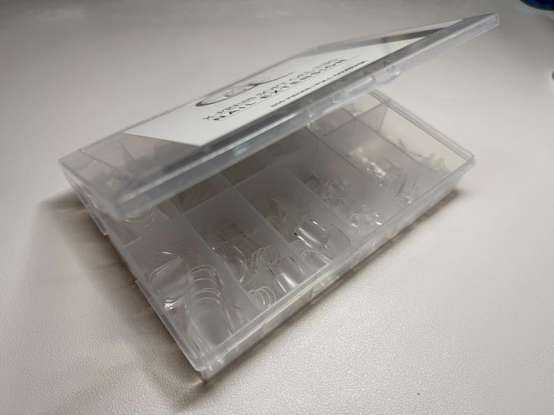 May include: A clear plastic box with a hinged lid, divided into multiple compartments. The box contains numerous clear, artificial nail tips in various sizes. The lid has a white label with black text. The box is open, revealing the contents.