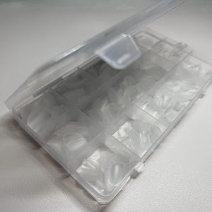 May include: A clear plastic storage box with a hinged lid, containing many translucent, oval-shaped nail tips. The box is divided into several compartments, each holding a different size of nail tips. The box is open, showing the contents.
