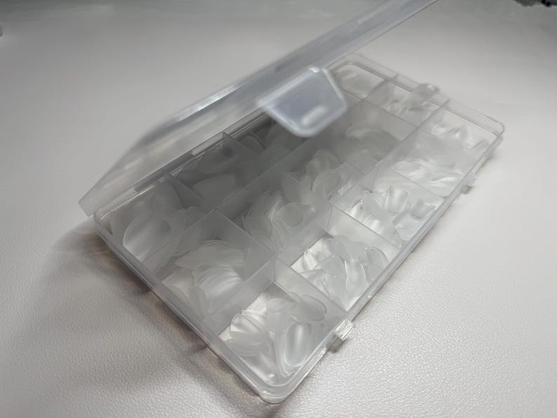May include: A clear plastic storage box with a hinged lid, containing many translucent, oval-shaped nail tips. The box is divided into several compartments, each holding a different size of nail tips. The box is open, showing the contents.