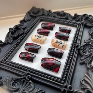 May include: A set of ten fake nails with a black and red blood splatter design. The nails have a white accent with a black handprint and the text "We Eat" on them.