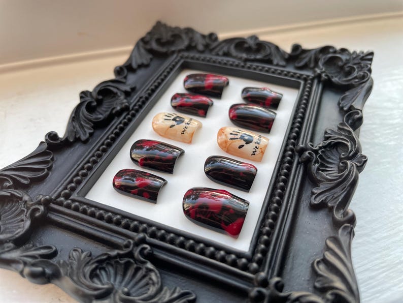May include: A set of ten fake nails with a black and red blood splatter design. The nails have a white accent with a black handprint and the text "We Eat" on them.