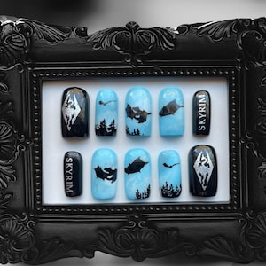 May include: A set of ten fake nails with a blue and black design. The nails feature a dragon silhouette, a mountain range, and the word "Skyrim" written in silver lettering.