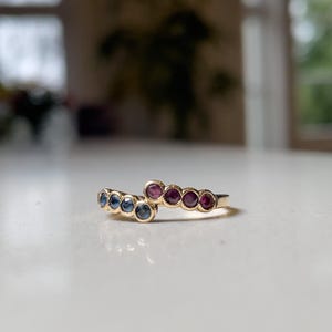 May include: Two gold rings with a curved design. One ring features a series of blue gemstones, while the other showcases a row of red gemstones. The rings are set on a white surface, with a blurred background.