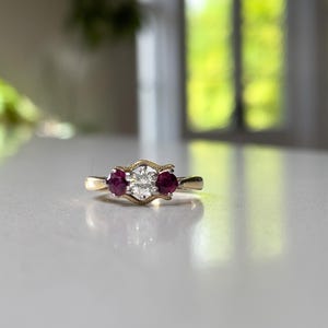 May include: A gold ring featuring a central round brilliant-cut diamond flanked by two round-cut red rubies. The setting has a textured, wavy metal design surrounding the stones, with a smooth, polished gold band completing the piece.