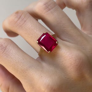 May include: A gold ring featuring a large, rectangular, faceted deep red gemstone set in a four-prong mounting. The stone is positioned centrally on the band, showcasing a vibrant crimson hue and clear, reflective surfaces across its top facets.