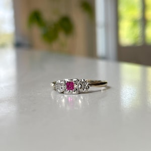 May include: A gold ring with a square, bright pink center stone, flanked by two smaller, round, clear stones. The ring's setting is silver-colored, and the band is gold. The ring is set against a white surface.
