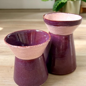 May include: Two handmade ceramic vases. The vases have a deep purple base and a light pink rim. The vases are on a wooden surface. The vases are of different sizes and shapes.