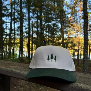 Può includere: Un cappellino da baseball con pannello frontale bianco e visiera verde scuro. Il pannello frontale presenta un ricamo di tre alberi di pino verde scuro disposti in fila. Il cappellino ha una corona strutturata e una visiera curva.