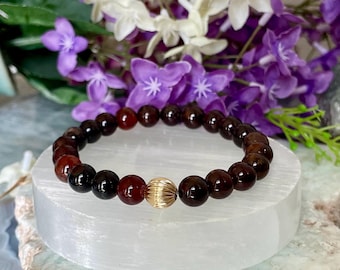 May include: A beaded bracelet featuring dark red and black beads with a gold-colored accent bead. The bracelet is displayed on a white surface, with purple and white flowers in the background. A jewelry item.