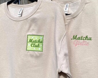 May include: Two tan sweatshirts with crew necks hang on black hangers. One sweatshirt has a green and white embroidered patch that says "Matcha Club." The other has "Matcha Girlie" embroidered in green and pink.