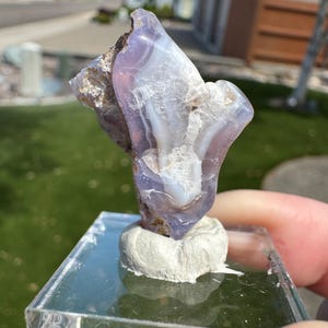 May include: A translucent lavender and white agate specimen with visible banding and crystalline textures, mounted upright on a white adhesive base attached to a clear, square acrylic display block for viewing.