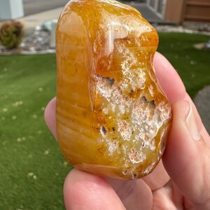 May include: A polished, translucent amber-colored stone with a smooth, rounded surface and a textured, crystalline patch featuring white and dark brown mineral inclusions. The stone exhibits a warm, golden-orange hue with subtle internal banding.
