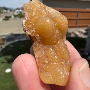 May include: A translucent, golden-orange botryoidal mineral specimen with a smooth, rounded, and bubbly surface texture. The stone exhibits varying shades of amber and honey tones, with a glossy, reflective finish across its irregular, organic shape.