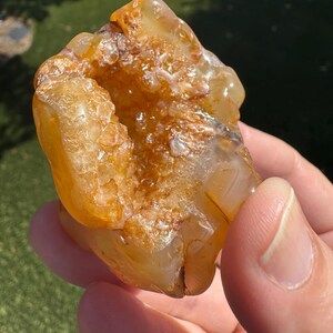 May include: A raw, translucent orange agate geode featuring a central cavity filled with sparkling, crystalline formations. The stone displays varying shades of amber and gold, with a smooth, polished outer surface contrasting against the rough, textured interior.