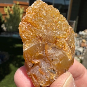 May include: A rough, amber-colored gemstone with a textured surface. The stone has a translucent quality, with variations in color and opacity. The natural rock formation is held in a hand, showcasing its organic shape and details.