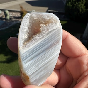 May include: An oval-shaped agate geode, polished to a smooth finish, displaying layers of white and grey. The geode's interior reveals a cavity lined with sparkling crystals. The stone is held in a hand, highlighting its natural patterns.