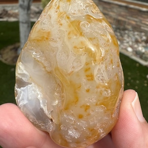 May include: A polished, teardrop-shaped agate stone featuring a translucent, milky white base with swirling patterns of golden-yellow and amber inclusions. The surface displays a smooth, glossy texture with natural crystalline pockets and crevices.