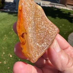 May include: A translucent, irregular piece of raw amber with a warm golden-orange hue. The surface displays a mix of smooth, polished sections and textured, matte areas with visible internal inclusions and deep orange streaks throughout.